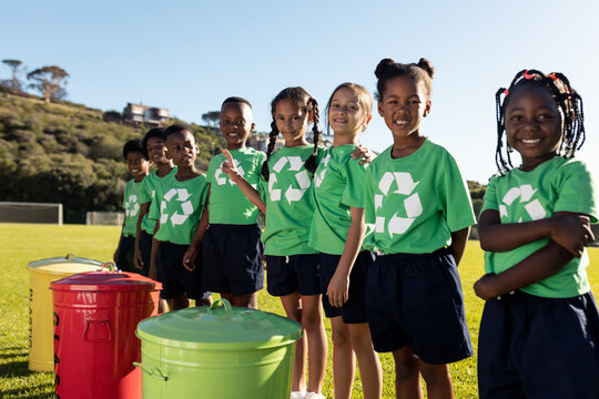 Portrait of happy diverse schoolchildren wearing green recycling tshirts at recycling bins - Powered by Adobe