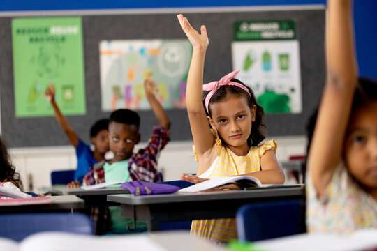 Diverse elementary schoolchildren sitting at desks and raising hands in elementary school class