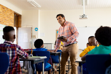Diverse male teacher with tablet and elementary schoolchildren sitting at desks in class
