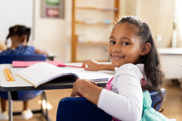 Portrait of happy biracial elementary schoolgirl sitting at desk in class