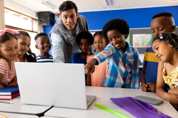 Diverse male teacher using laptop teaching children in elementary school class