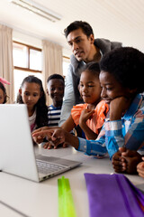 Diverse male teacher using laptop teaching children in class at elementary school