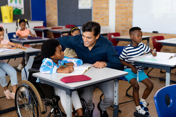 Happy biracial male teacher with boy in wheelchair in class at elementary school