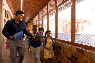 Diverse male teacher and children walking together in corridor at elementary school, copy space