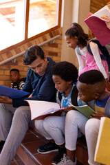 Happy diverse male teacher and children with books sitting on stairs in class at elementary school