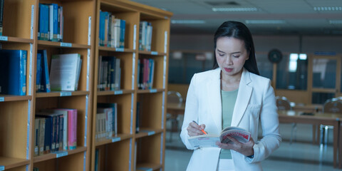A female university lecturer in a business suit is searching for the book on the bookshelf in the...