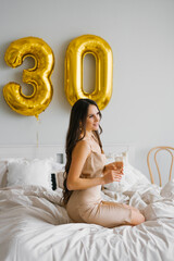 Obraz na płótnie Canvas Long haired birthday woman sitting on the bed in the morning and holds a glass of champagne in her hands and smiles celebrating her thirtieth birthday