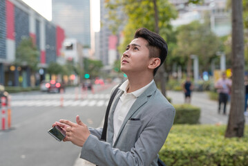 A young asian man looking up to a tall building, confirming an address on his gps mobile app. Using a map with location ping on his cellphone.