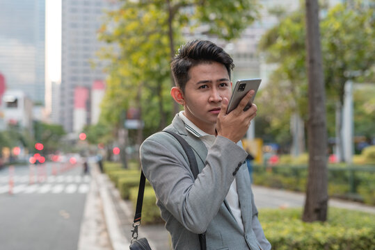 A young man talks to someone via speakerphone or using voice recognition app technology to get directions to an address on his cellphone while walking. Outdoor city scene.