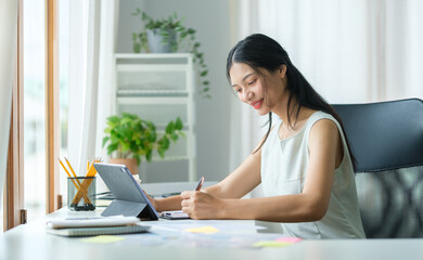 Young asian female office worker sitting in bright office and working on digital tablet.