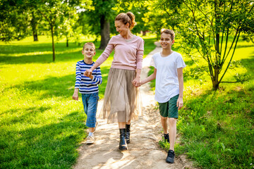 Fototapeta premium Happy mother is walking with her sons in park. They are holding hands and enjoying summer day.
