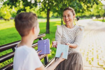 Naklejka premium Happy mother is sitting with her son on bench in park. Boy is giving a present to his mother.