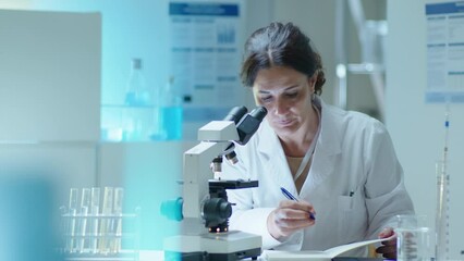 Mature female scientist in white coat using compound microscope and making notes, conducting chemistry research in laboratory - Powered by Adobe