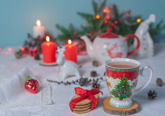 A cup of tea with a Christmas pattern on a table with a linen tablecloth, burning candles, a teapot and fir branches.