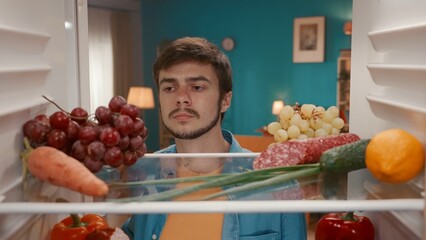 A young frustrated man looks inside the refrigerator. A man examines healthy products on the...