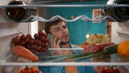 A man with a phone to his ear at the open refrigerator. A man inspects the food in the refrigerator, and makes an order for delivery. Purchase, order foods. View from inside the refrigerator.