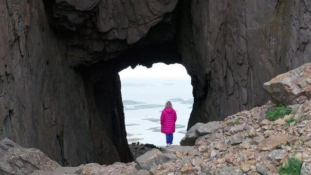 Blonde in pink jacket by hole through Torghatten mountain; drone crane reveal