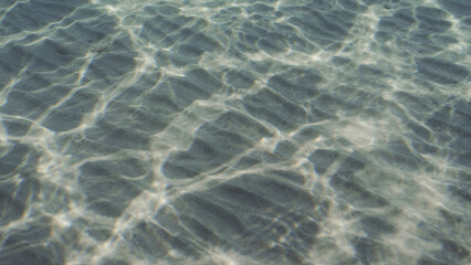 Fototapeta na wymiar Glare of sun plays on sandy bottom in shallow water. Top view on sandy seabed in shallow water with diagonal lines of sand and sun glare on its surface in brightly suny day, Red sea, Egypt