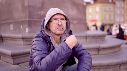Portrait of adult man with glasses sitting in the hood on square and smoking a tobacco pipe in the Palace Square, Warsaw Old Town