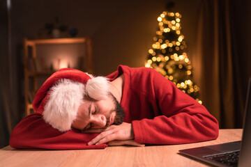 Exhausted young businessman in Santa hat sleeping by desk in front of laptop on Christmas night in home office