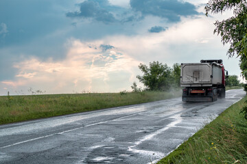 Heavy truck vehicle driving wet asphalt road driveway in stormy rainy weather with epic clouds and green landscape. Rear car view with water mist on wheels