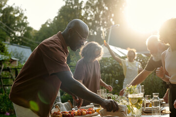 Side view of intercultural family putting homemade food and drinks on table for dinner while preparing for meeting guests or outdoor gathering