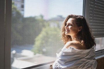 Young smiling caucasian woman in a white shirt standing on the balcony in the morning meeting a new...