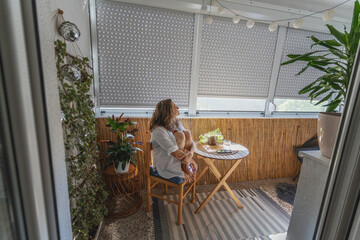 Young caucasian woman in a white shirt sitting on a balcony with roller shutters. Sun protection and rising temperature concept