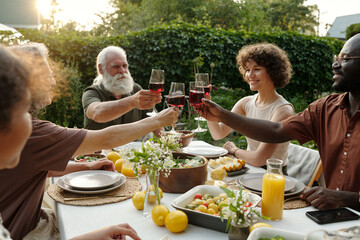 Happy intercultural members of large family toasting with glasses of red wine while sitting by served table outside and celebrating holiday