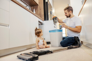 A man is cleaning vacuum cleaner and emptying it in a garbage can while his baby is looking at him.