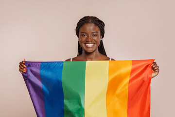 Young cheerful african girl with braids holds up rainbow flag at studio isolated over beige background. Gay pride. She fights for sexual freedom. - obrazy, fototapety, plakaty