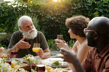 Senior man with white beard and hair holding glass og orange juice and talking to young intercultural couple by served table during family dinner