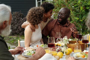 Happy cute African American boy embracing his intercultural parents sitting by table with homemade food and drinks during outdoor dinner
