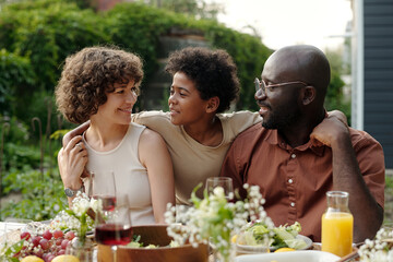 Cute little boy looking at his mother while embracing parents sitting by table served for outdoor family dinner and communicating to their son