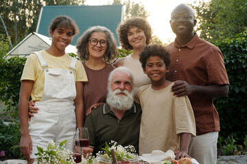 Happy large intercultural family of three generations looking at camera with smiles while standing in front of served table during outdoor dinner