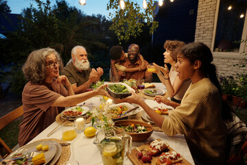 Cute girl taking container with steamed broccoli from hands of her grandmother over served table with homemade food and drinks