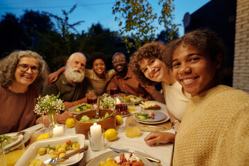 Happy African American girl taking selfie with large intercultural family sitting by table served with homemade food for outdoor dinner