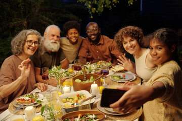 Large intercultural family looking at smartphone screen while communicating with their friends in video chat during outdoor dinner