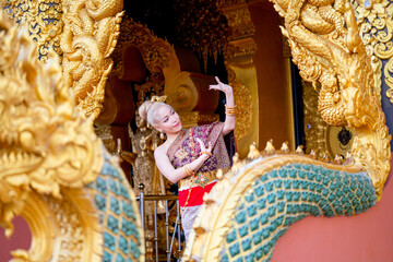 Pretty Asian woman wear Thai traditional dress stand and action of Thai dance in front of door of temple and naga sculpture as foreground.