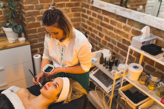 Cosmetologist applying charcoal facial mask using a brush to her client in a beauty spa. High quality photo