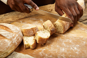 Close up of african-american man slices fresh crispy bread with kitchen knife on wooden table.