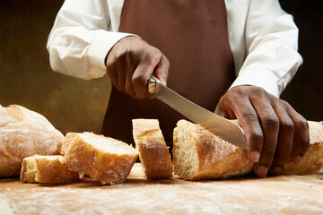Image of african american man slices fresh crispy french baguette with knife on wooden table.