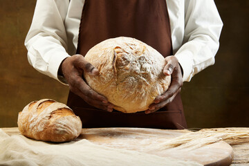 Unrecognizable african baker in brown apron holding round bread in hands and next to board where...