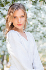 Portrait of a young attractive woman in a white dress against the backdrop of blooming apple trees. The girl poses against the background of flowers in the spring park.