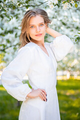 Portrait of a young attractive woman in a white dress against the backdrop of blooming apple trees. The girl poses against the background of flowers in the spring park.