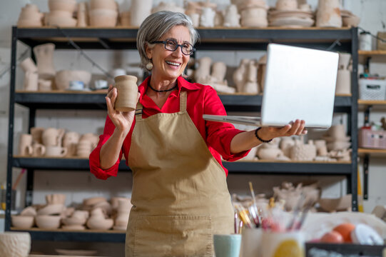 Woman pottery artist using laptop in art studio showing her earthenware products, selling online.
