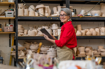 Female potter with tablet computer in art studio among earthenware products on shelves.