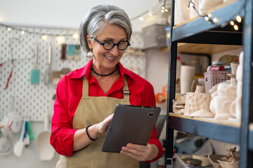 Happy female ceramist using digital tablet in store, managing store with handmade ceramic products