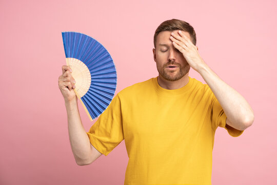 Too hot. Sweaty tired middle aged man touching forehead using paper fan suffer from heat, feels sluggish. Displeased guy cooling in hot summer weather, isolated on studio pink background. Overheating