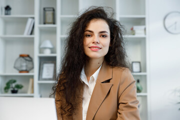 Young curly brunette girl with a laptop looks at the camera. Freelancer portrait working from home, remote work. Successful manager girl, in a white office. Reports online.